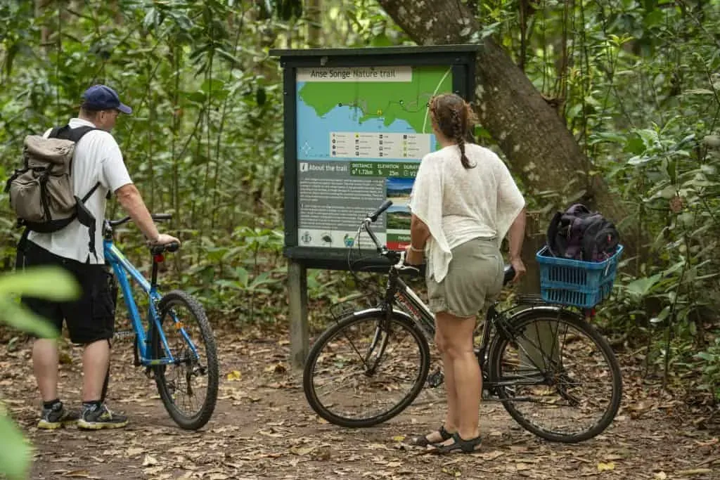 Two cyclists reading an informational board about the Anse Songe Nature Trail surrounded by lush tropical forest in Seychelles.