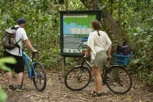 Two cyclists reading an informational board about the Anse Songe Nature Trail surrounded by lush tropical forest in Seychelles.