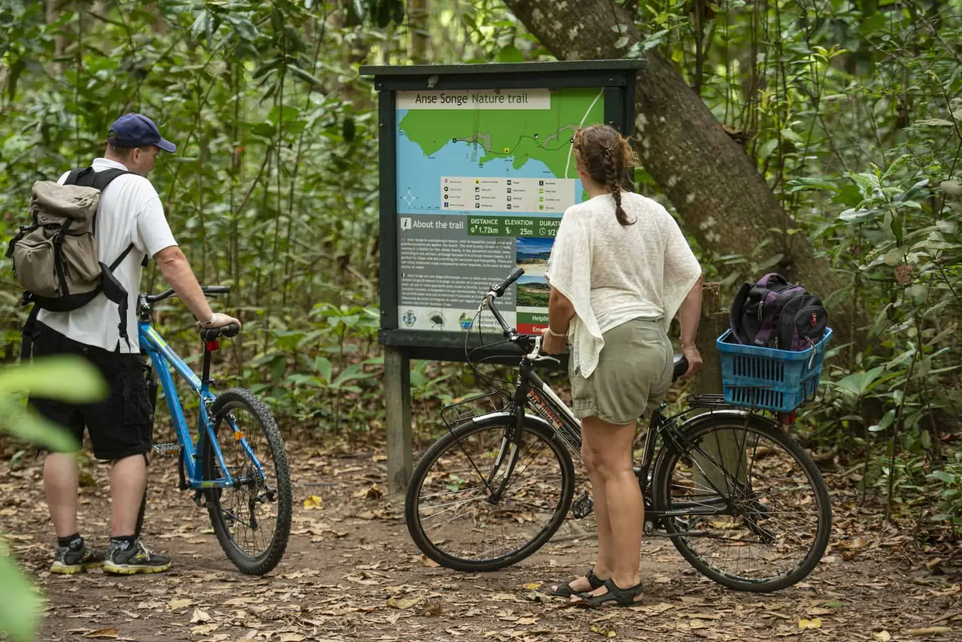 Two cyclists reading an informational board about the Anse Songe Nature Trail surrounded by lush tropical forest in Seychelles.
