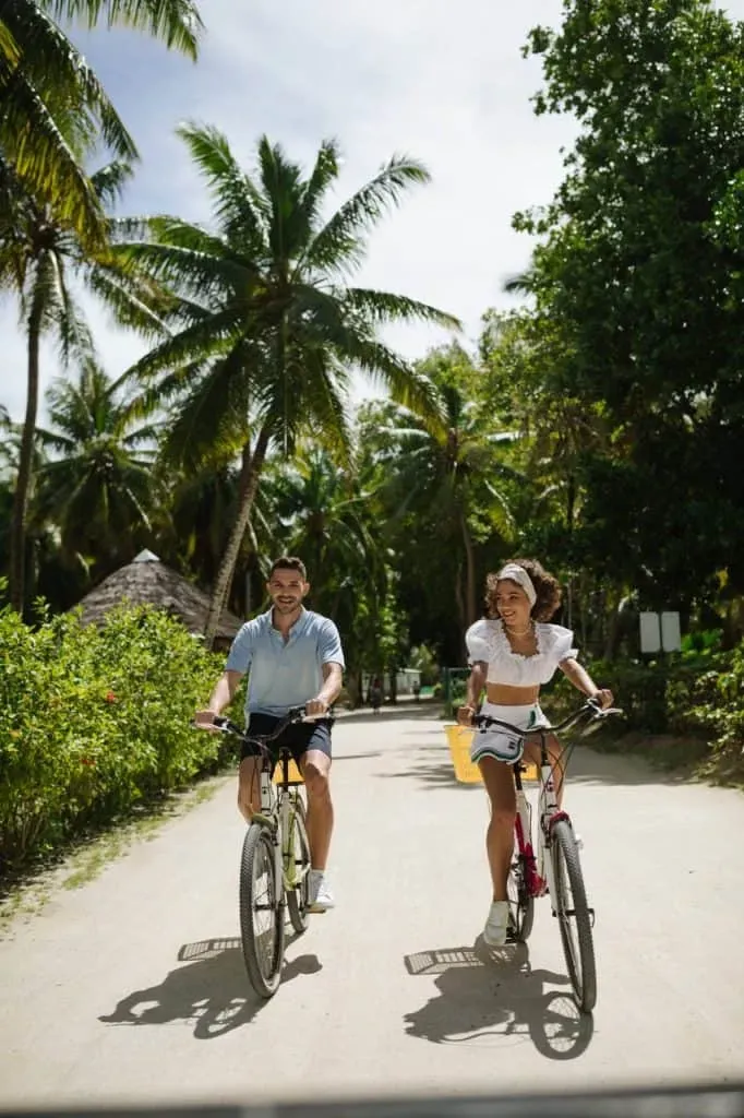 Two people cycling along a sandy path surrounded by palm trees and tropical greenery on La Digue Island, Seychelles.