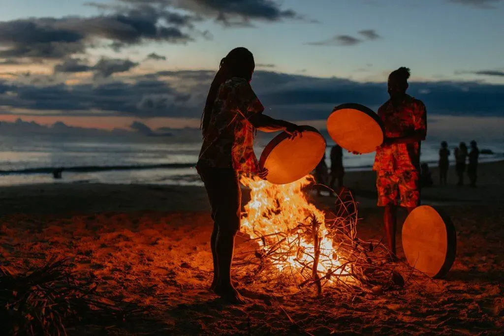 Two men performing a drum ritual around a bonfire on the beach at sunset in Seychelles, with silhouettes and glowing flames.
