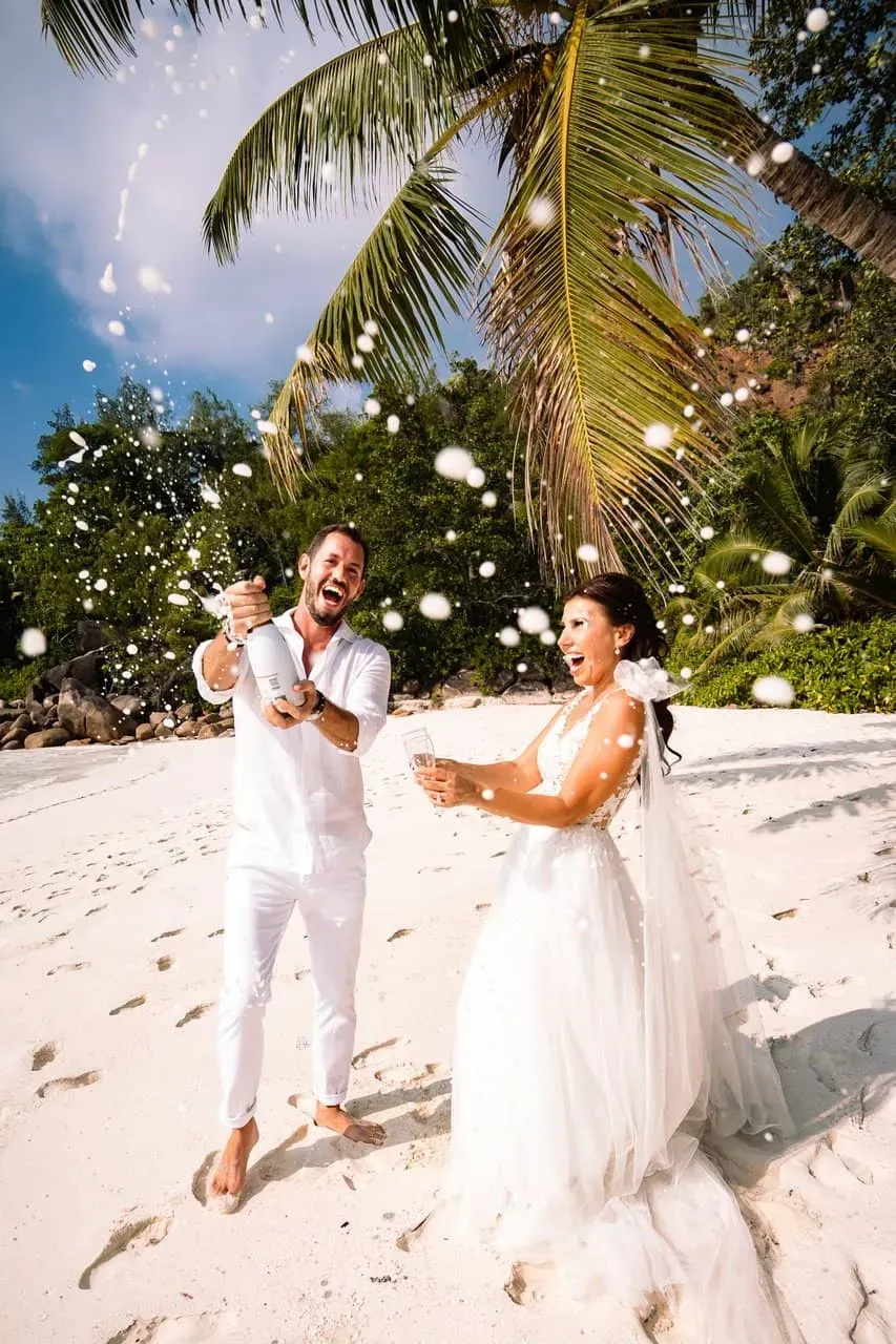 A bride and groom celebrating their wedding on a tropical beach, with the groom popping a champagne bottle under a palm tree.