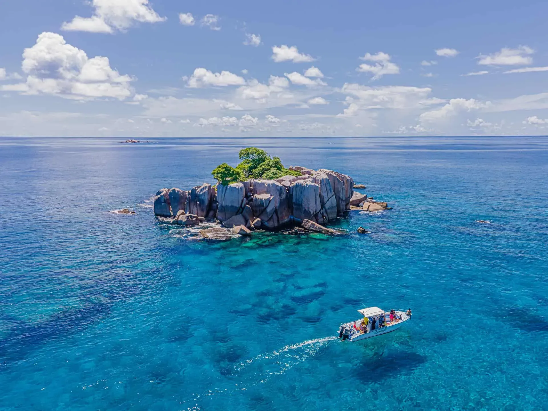 A motorboat approaching a secluded granite islet surrounded by turquoise waters under a bright blue sky in Seychelles.