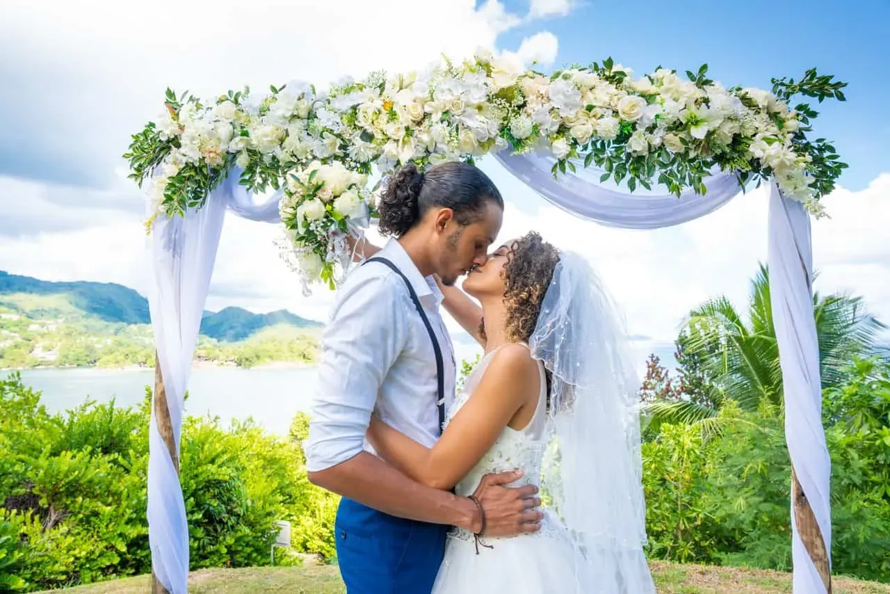 A bride and groom sharing a kiss under a floral wedding arch with lush greenery and ocean views in Seychelles.