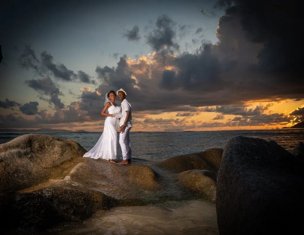 A bride and groom standing on granite rocks by the ocean during sunset in Seychelles, with dramatic clouds and golden hues in the sky.