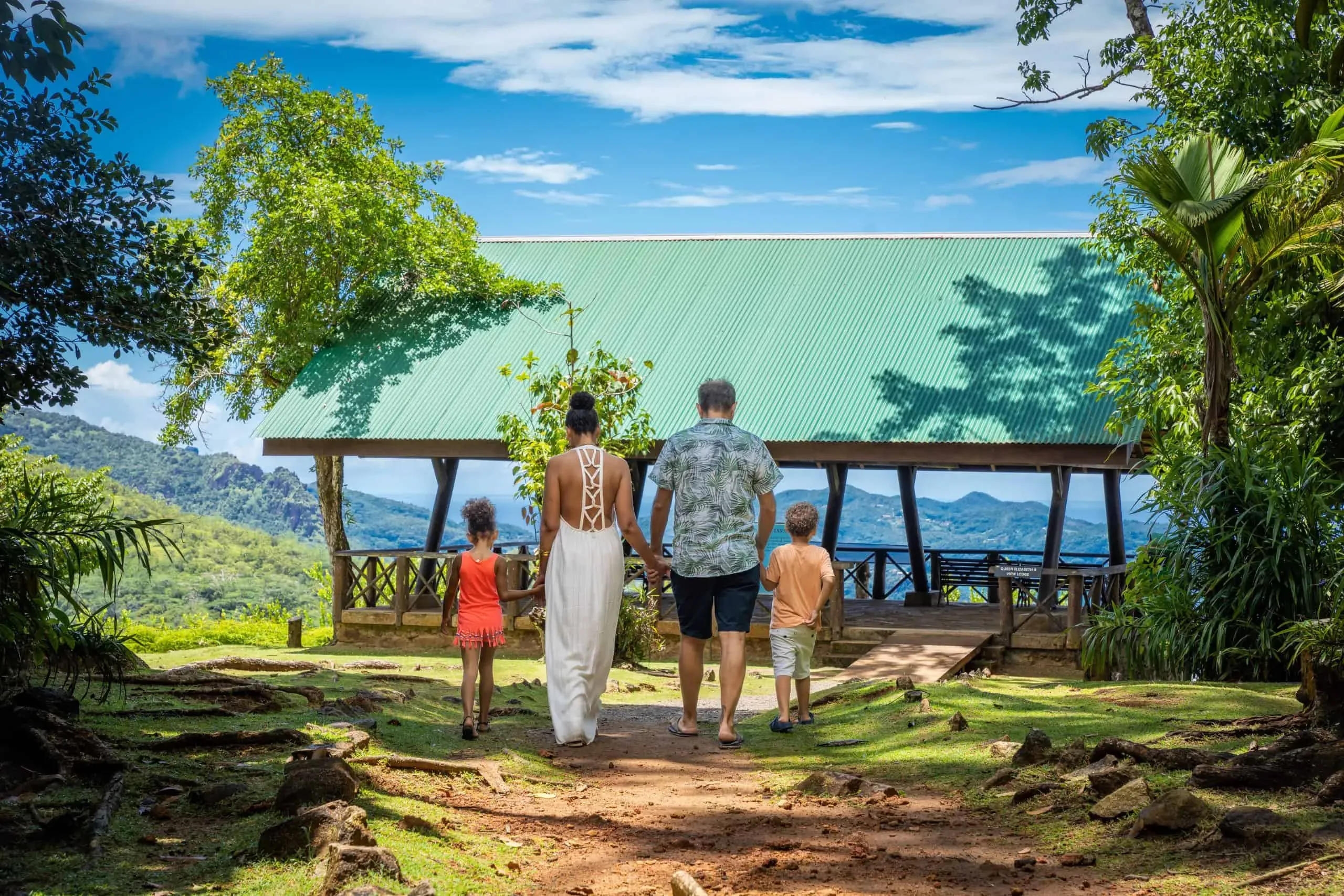 A family walking toward a green-roofed pavilion overlooking mountain vistas and lush landscapes in Seychelles.
