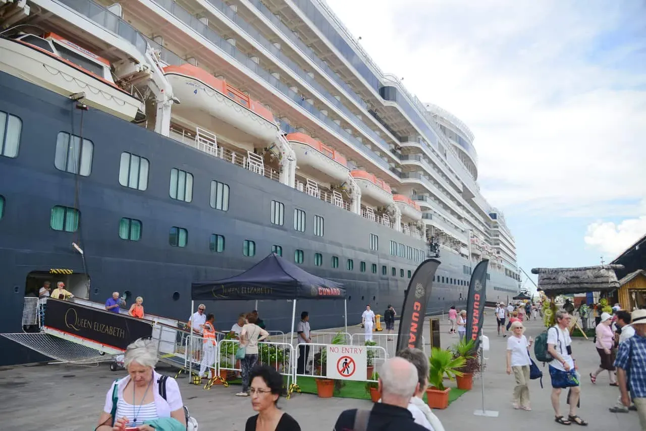 The Queen Elizabeth cruise ship docked at a Seychelles port with passengers disembarking and exploring the harbor area.