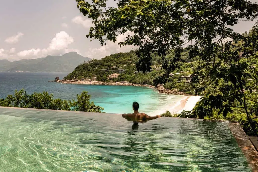 A man enjoying an infinity pool with breathtaking views of turquoise waters, white sandy beaches, and lush green hills in Four Seasons Resort Seychelles, Seychelles.