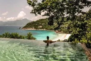 A man enjoying an infinity pool with breathtaking views of turquoise waters, white sandy beaches, and lush green hills in Four Seasons Resort Seychelles, Seychelles.