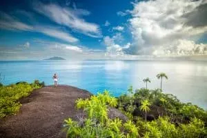 A woman standing on a granite rock overlooking the vast turquoise ocean and distant islands under a bright blue sky in Anse Major Trail, Seychelles.