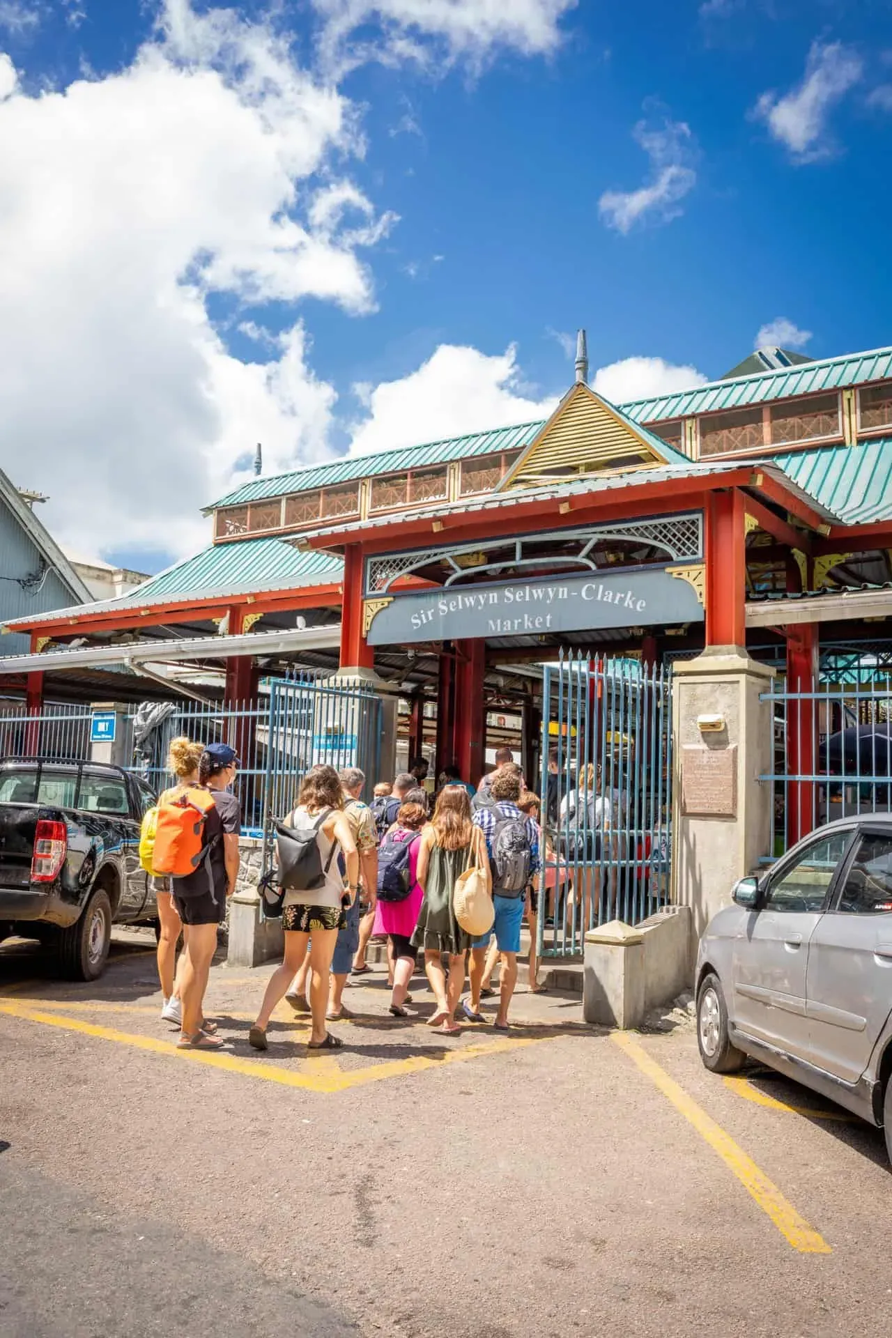 Tourists entering the historic Sir Selwyn Selwyn-Clarke Market in Victoria, Seychelles, with colorful architecture and blue sky overhead.