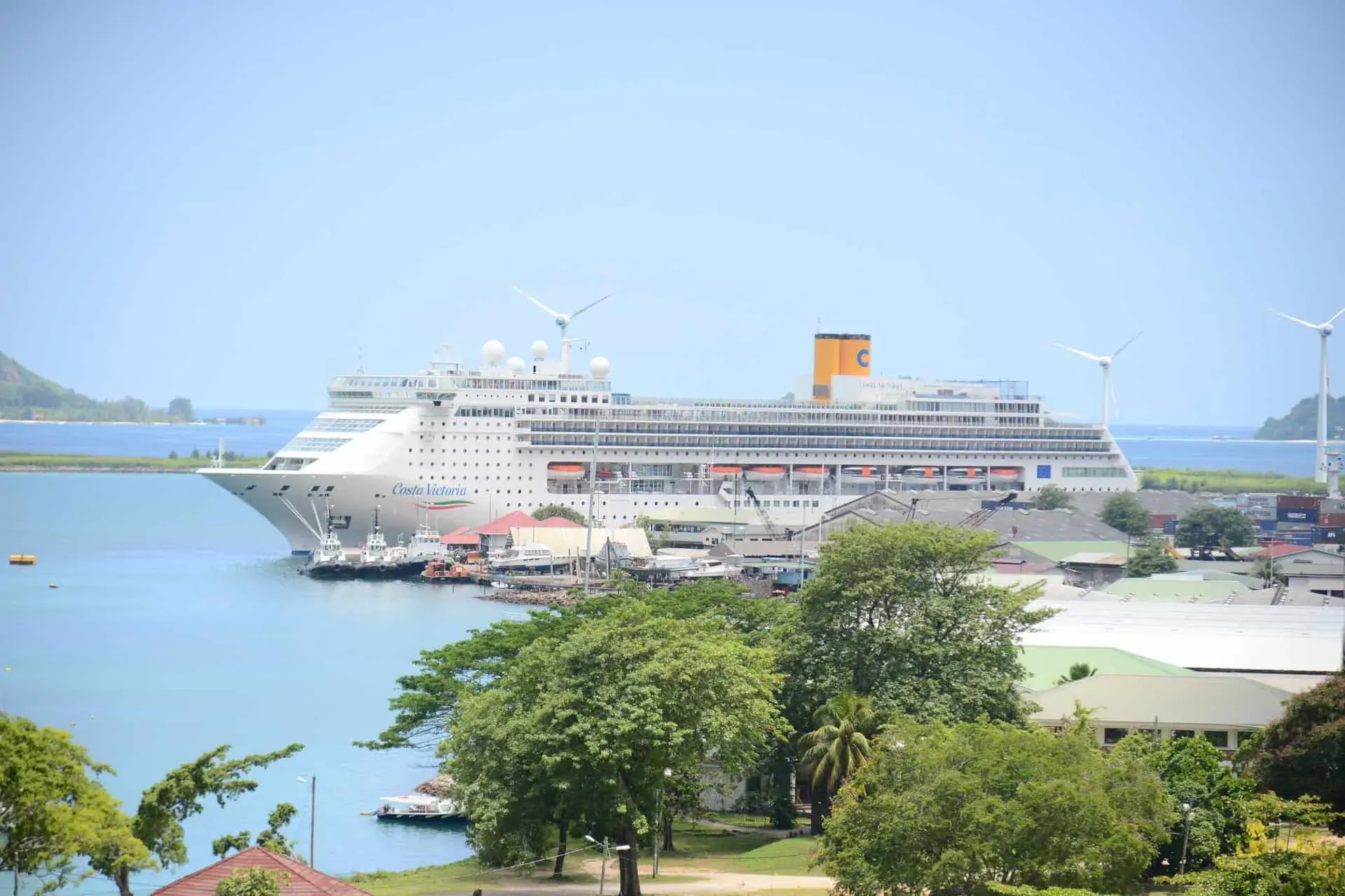 Cruise ship docked at Port of Victoria with surrounding port facilities, wind turbines, and lush greenery in Seychelles.