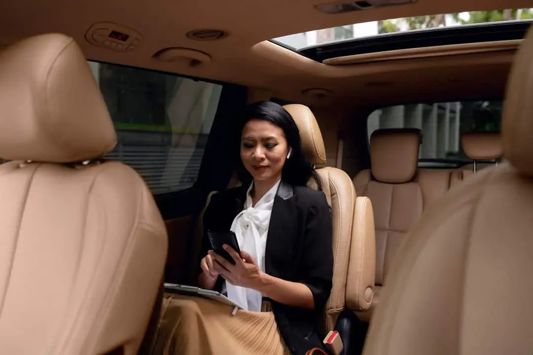 Businesswoman sitting comfortably in the backseat of a luxury car, using her mobile phone and tablet, during a VIP transfer service.