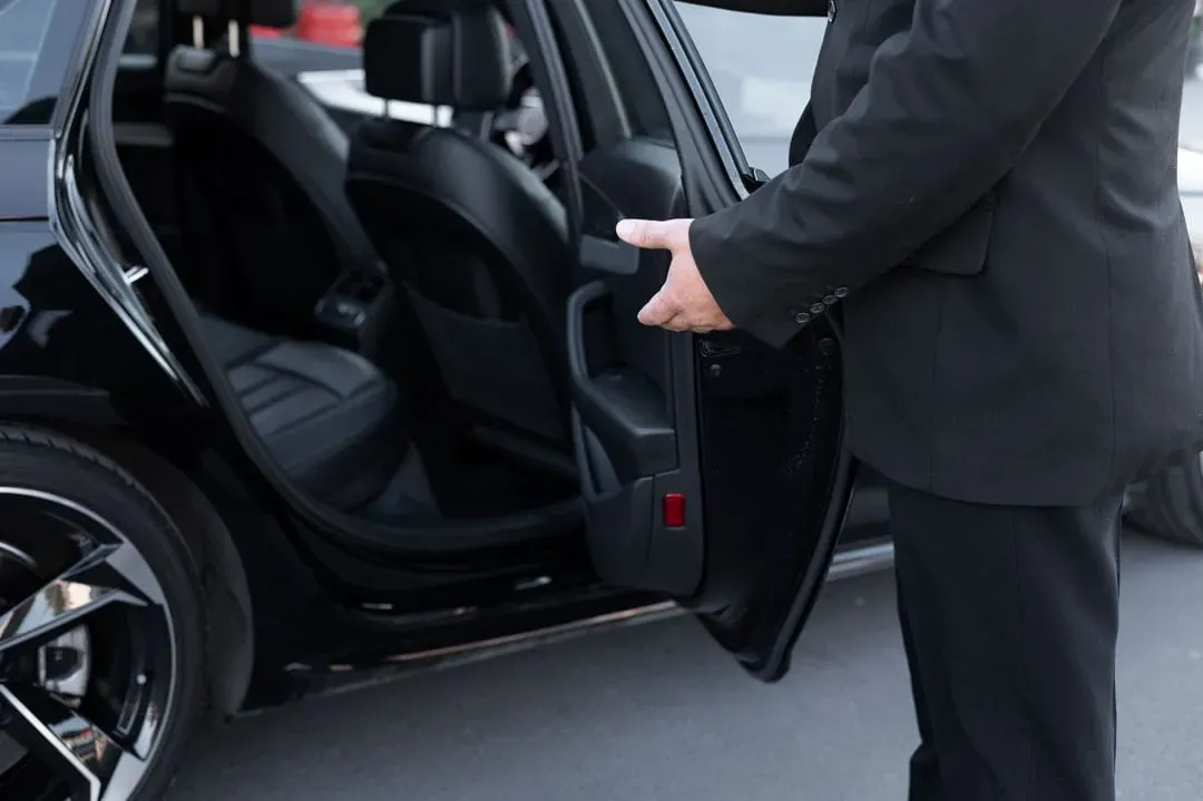 Chauffeur in a suit politely opening the rear door of a sleek black car ready for a VIP guest or client.