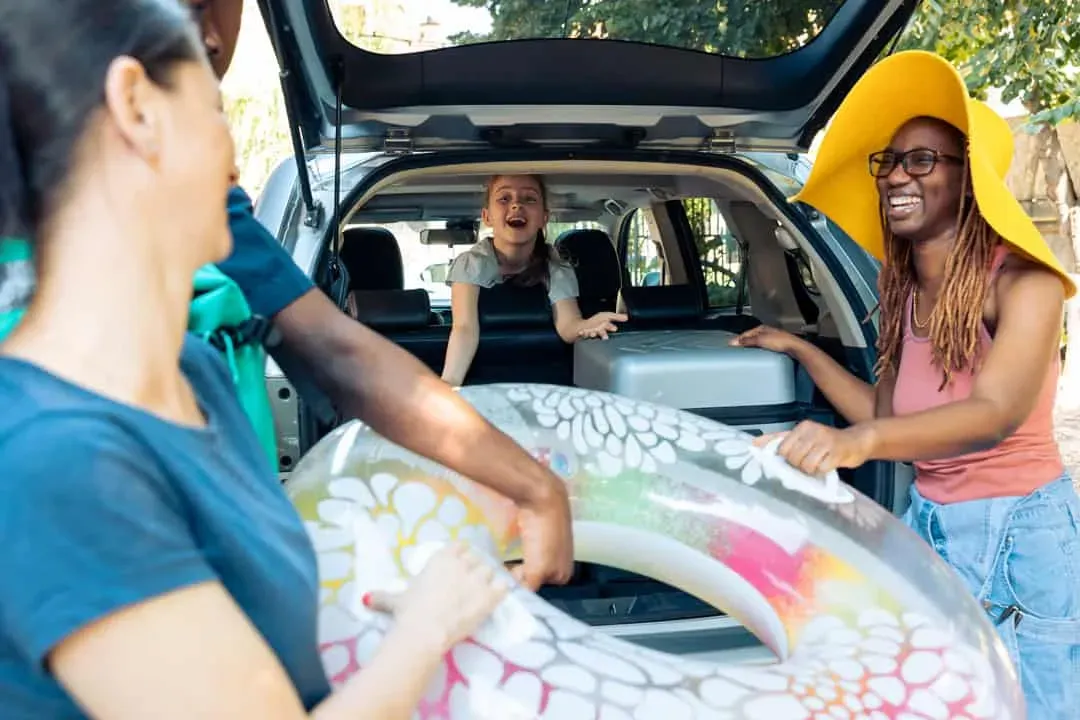 Group of cheerful friends and family loading bags and an inflatable float into the back of an SUV, preparing for a seaside vacation.
