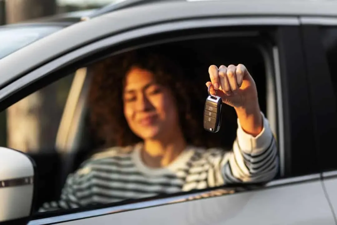 Young woman sitting in the driver’s seat inside a rented car, holding car keys up and smiling