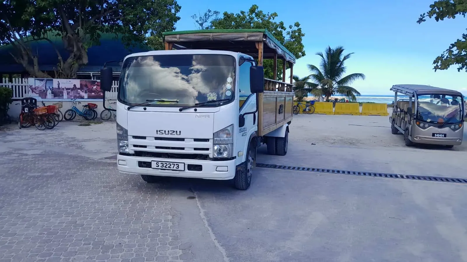 Isuzu utility trucks parked near a beach with bicycles and palm trees, providing transportation for Cheung Kong Travel in a tropical coastal location.