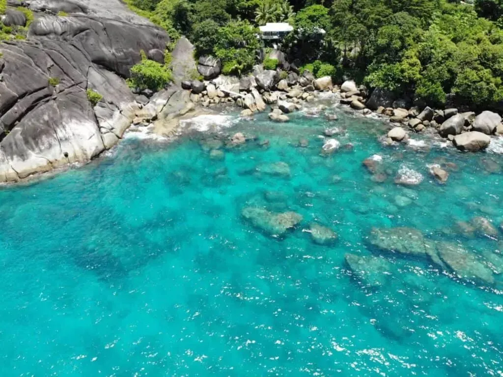 Aerial view of black granite cliffs and lush greenery beside brilliant turquoise water dotted with underwater rocks, Seychelles.