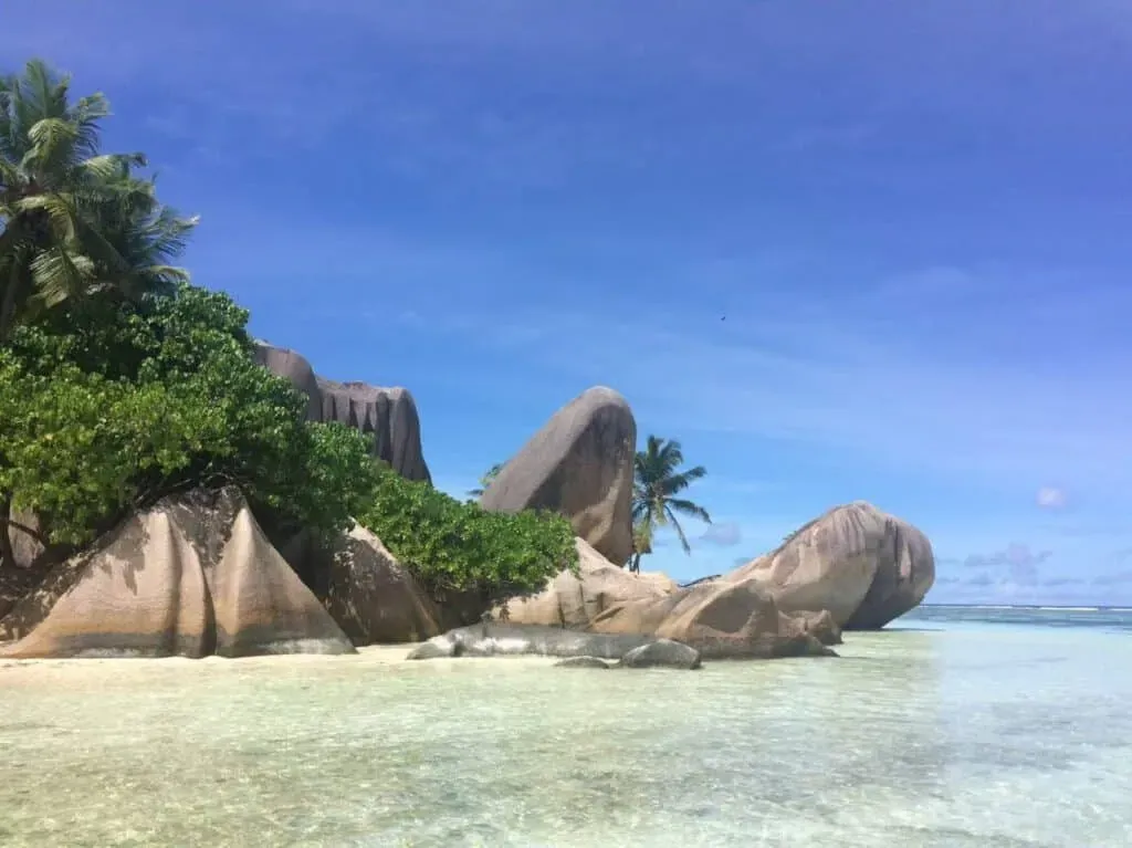Spectacular granite boulders and tropical greenery beside shallow, clear turquoise water under a vivid blue sky at Anse Source d’Argent, Seychelles.