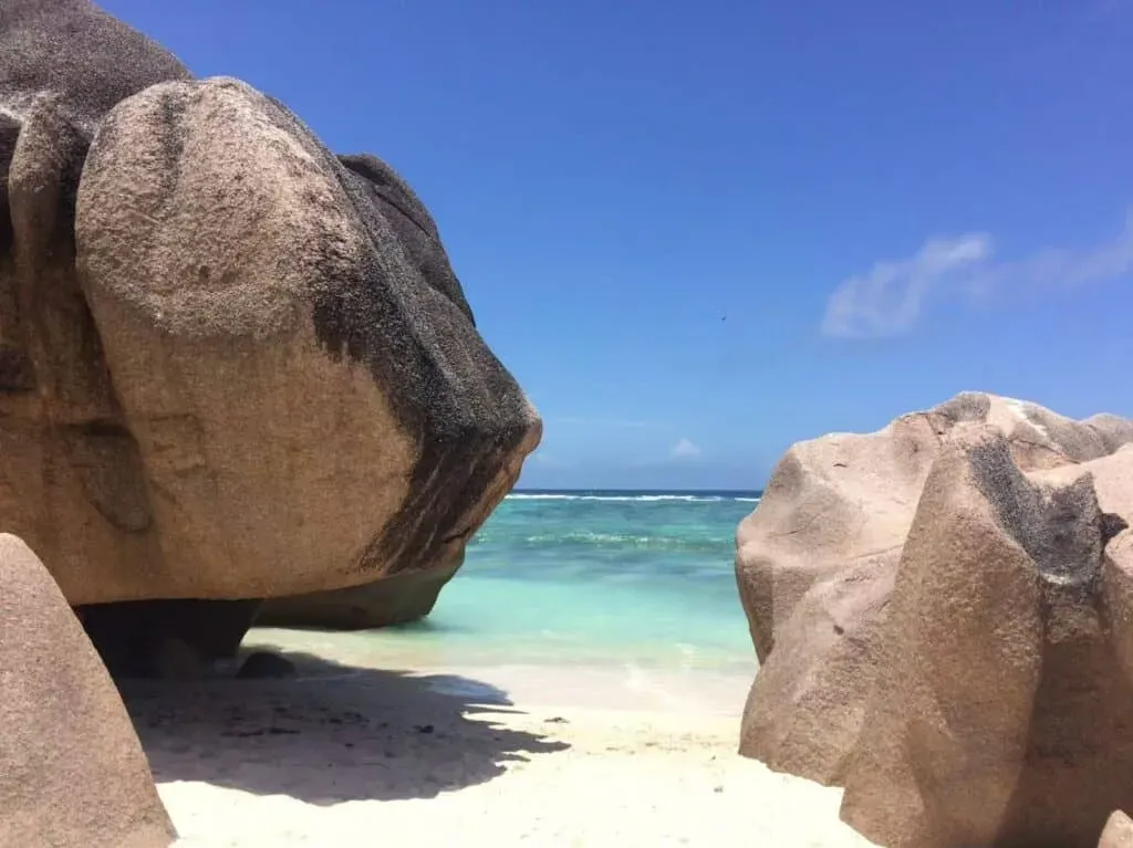 Giant granite boulders on a sunlit white sand beach by clear turquoise waves under a bright blue sky in Seychelles.