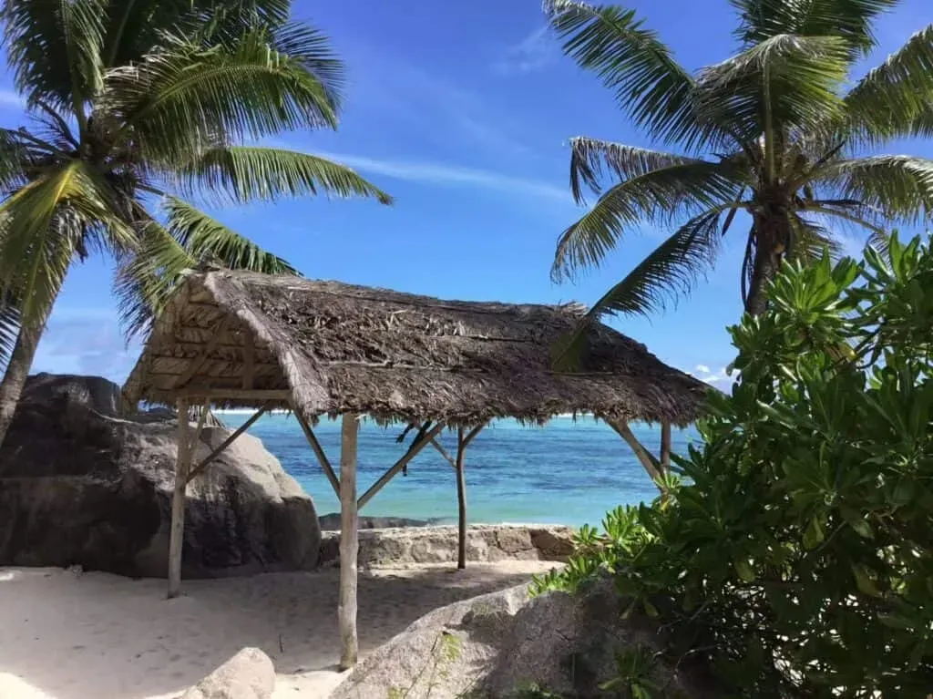 Thatched beach shelter surrounded by palm trees on a sandy shore, with the turquoise ocean and blue sky in the background in Seychelles.