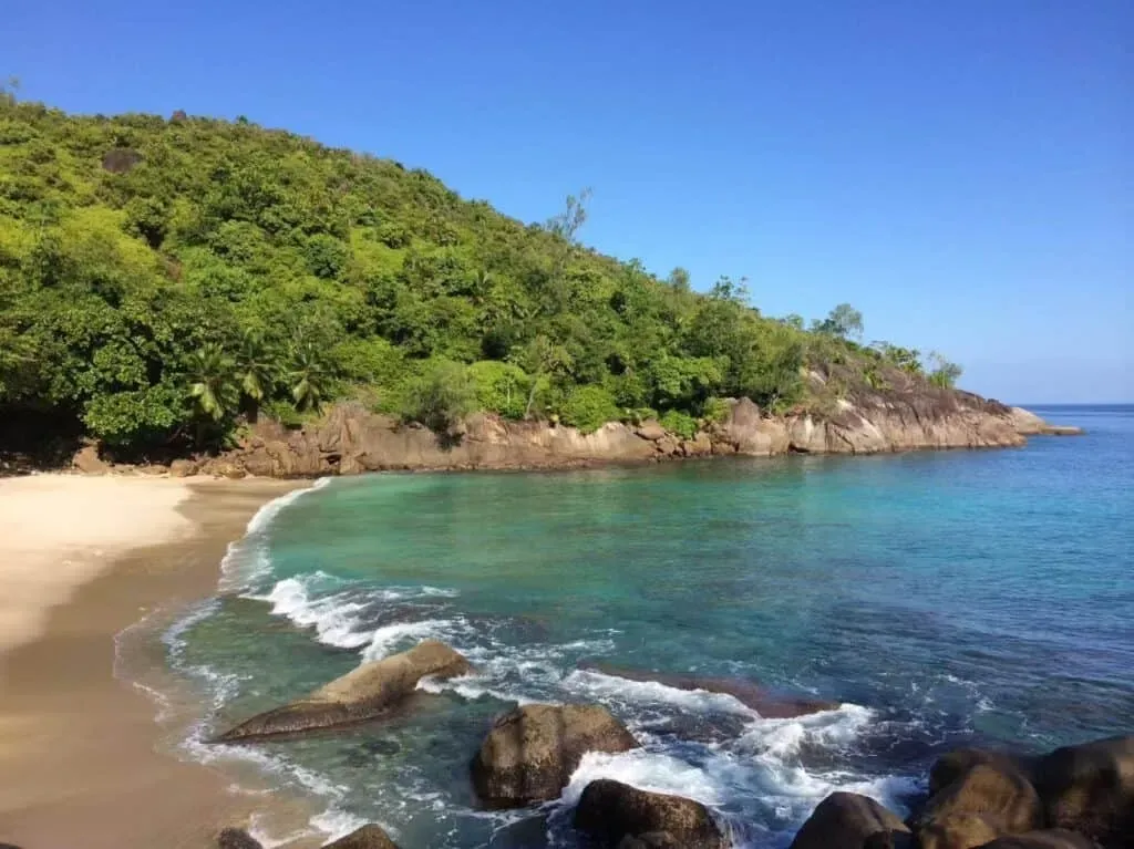 Rocky shoreline with turquoise waters, sandy beach, and lush green hillside under a clear blue sky in Seychelles.