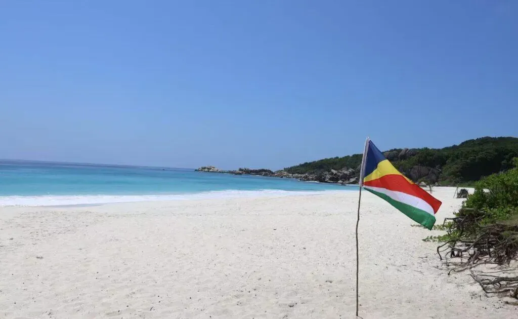 Seychelles national flag standing on a pristine sandy beach with turquoise waters and clear blue sky.