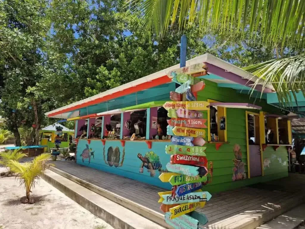 Colorful beach bar with travel-themed painted signs showing international destinations, surrounded by tropical trees in Seychelles.
