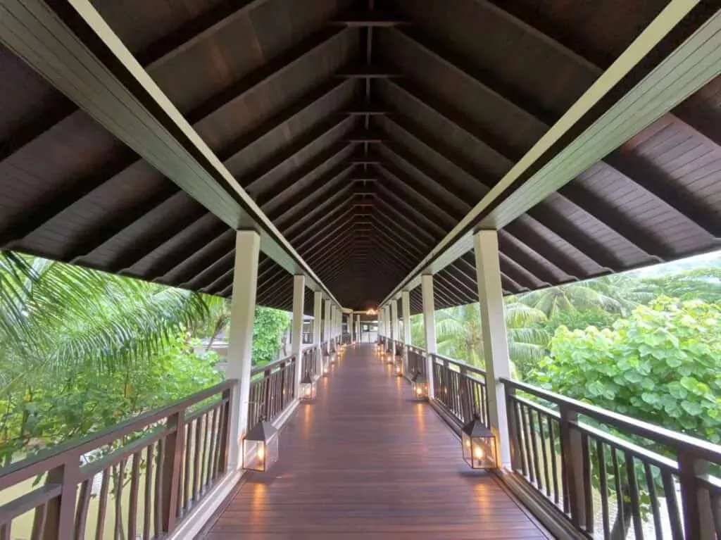 Covered wooden walkway with lanterns and railings, surrounded by tropical palm trees and lush greenery in Seychelles.