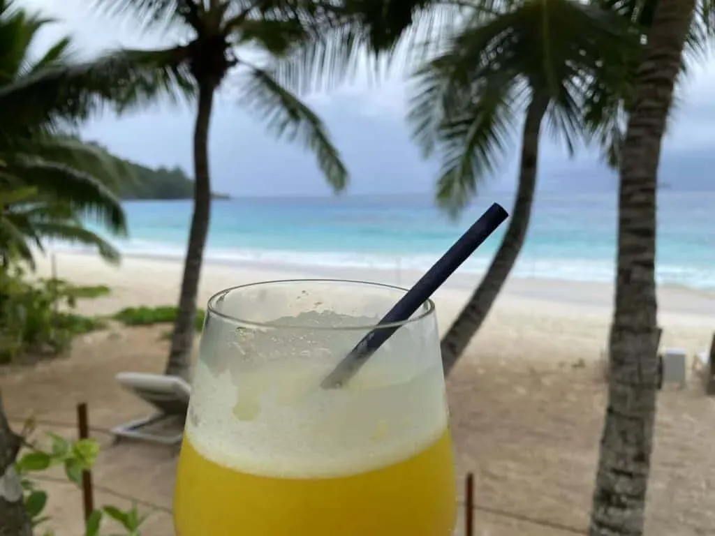 Fresh tropical juice in a glass with a straw, set against a sandy beach, palm trees, and turquoise ocean waves in Seychelles.