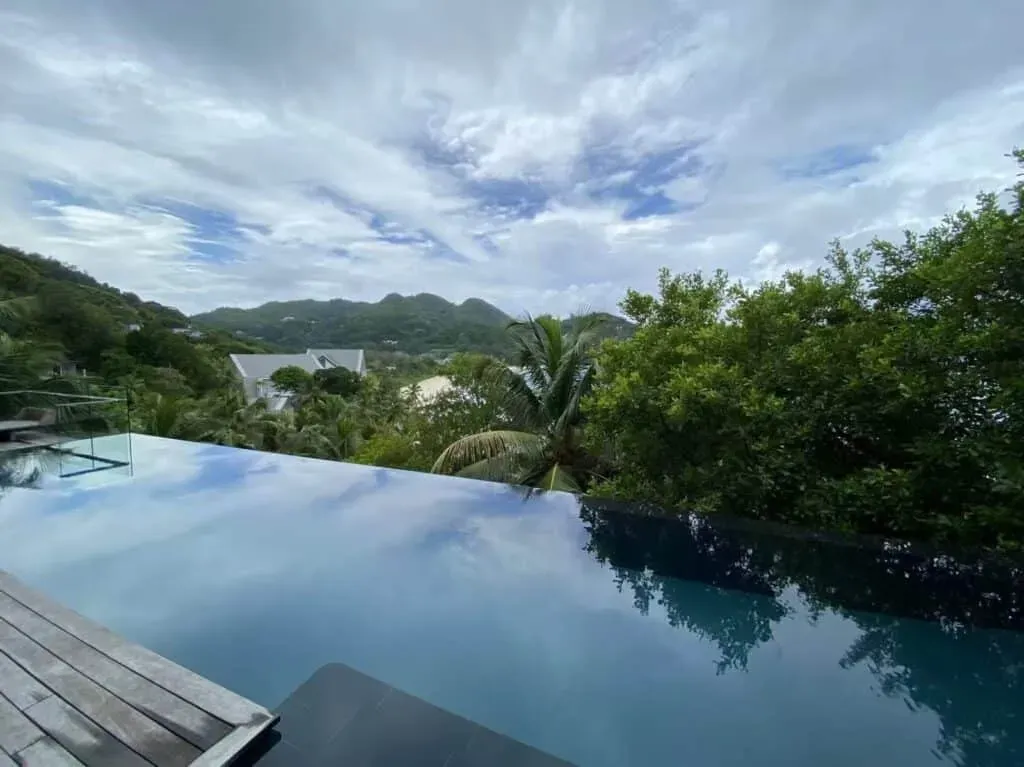 Infinity pool with blue water reflecting the sky, surrounded by tropical trees and hills in Seychelles.