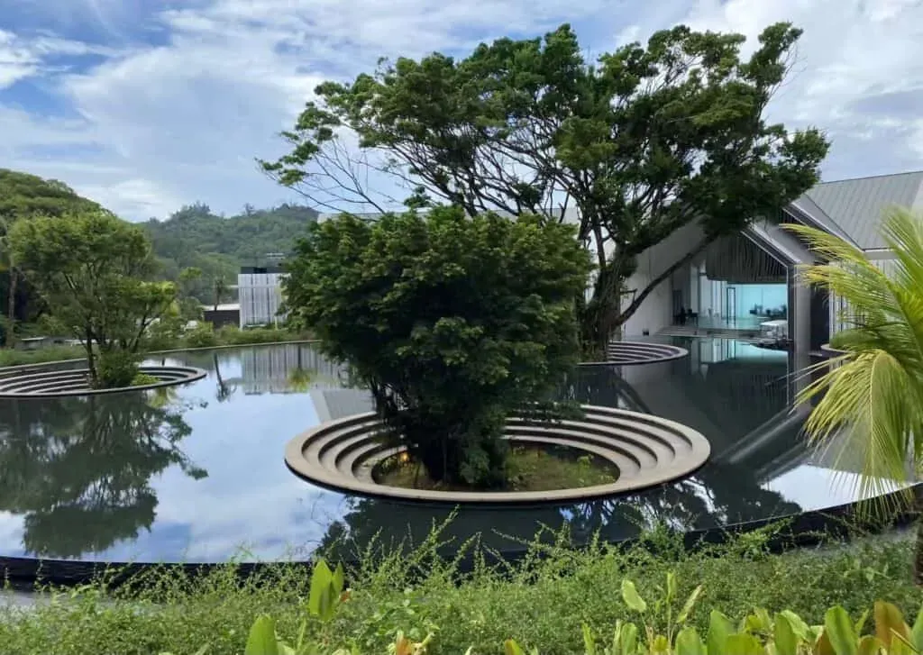 Contemporary water garden with circular sculptural platforms featuring lush green trees, reflecting a modern building and natural landscape in Seychelles.